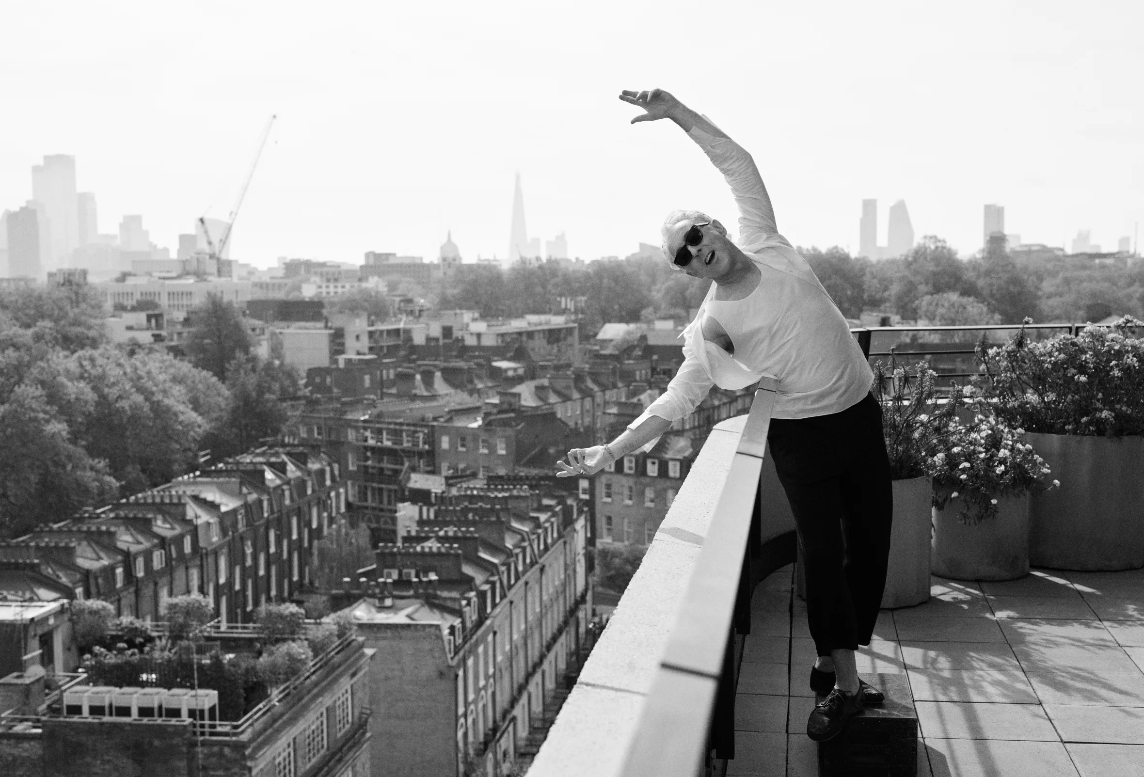 A grayscale photo of a man in sunglasses leaning over a rooftop railing with a city skyline in the background.