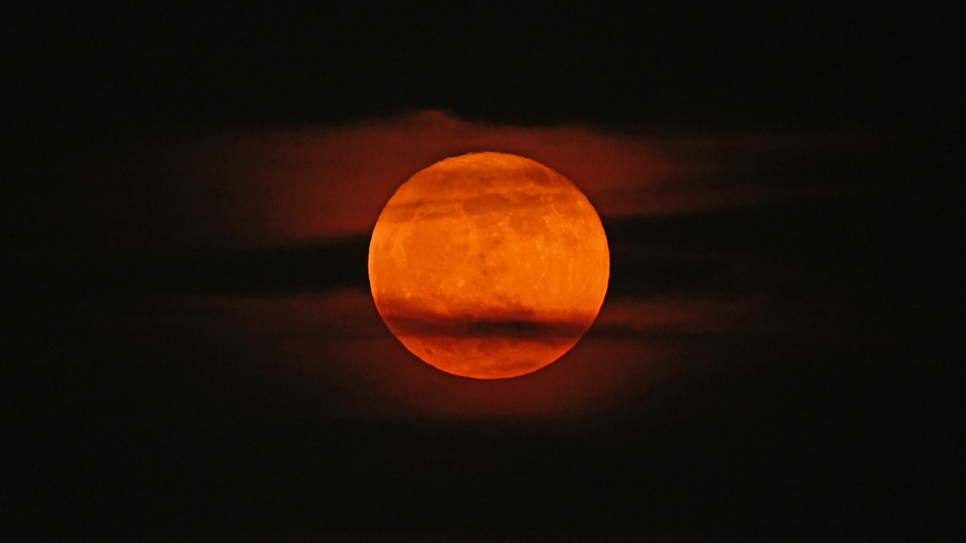An orange-red moon glows in a dark night sky with streaks of cloud passing across its surface.