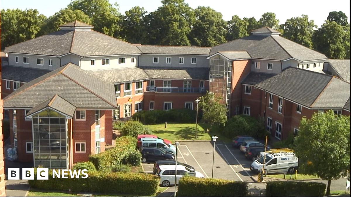 An aerial view of a large brick built hospital. There are cars parked in the car park at the front and the area is surrounded by trees with green leaves.