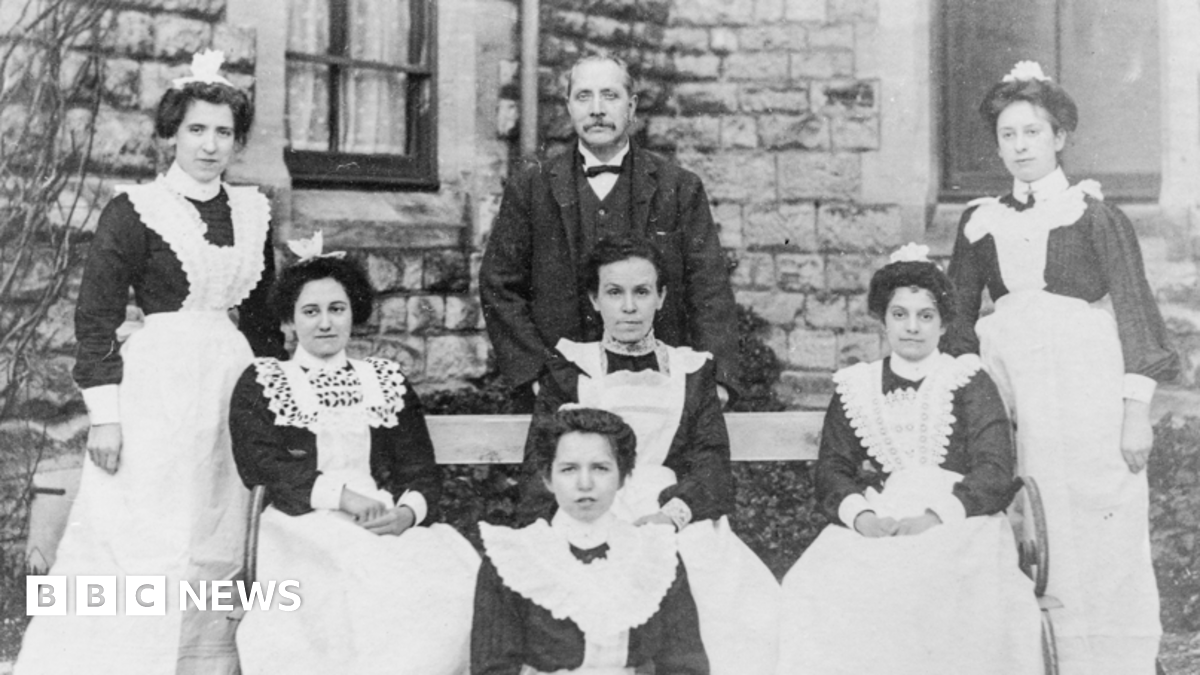 Three rows of women wearing Victorian hospital uniforms - dark dresses and white aprons - and a man are pictured sitting or standing in front of a stone building.