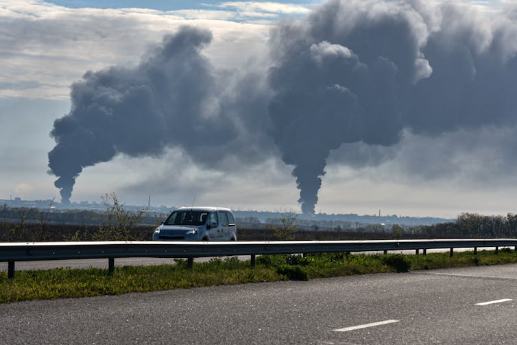 a car drives on a road, tall plumes of smoke rise in the background