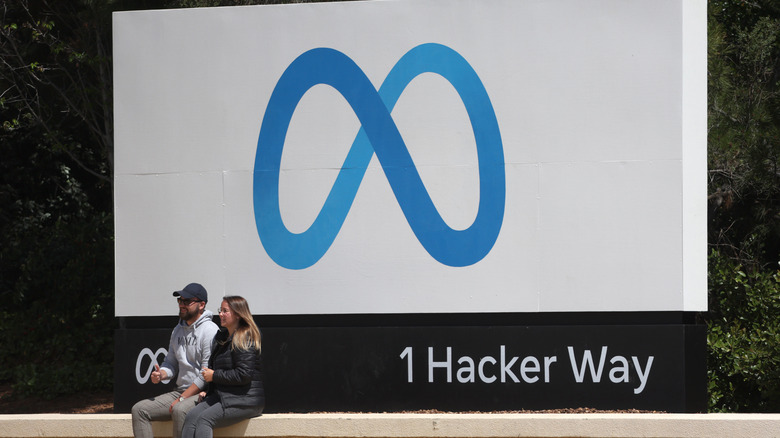 Two people in front of Meta's logo marker in its headquarters.