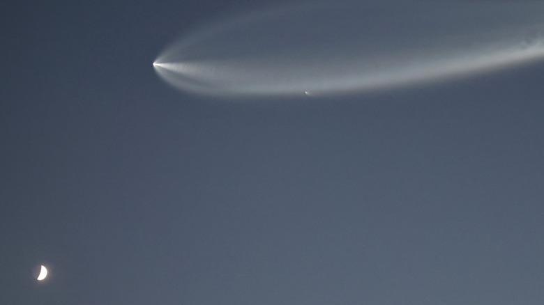 A Falcon 9 rocket disappears into the atmosphere leaving a wide plume behind it above a half-moon