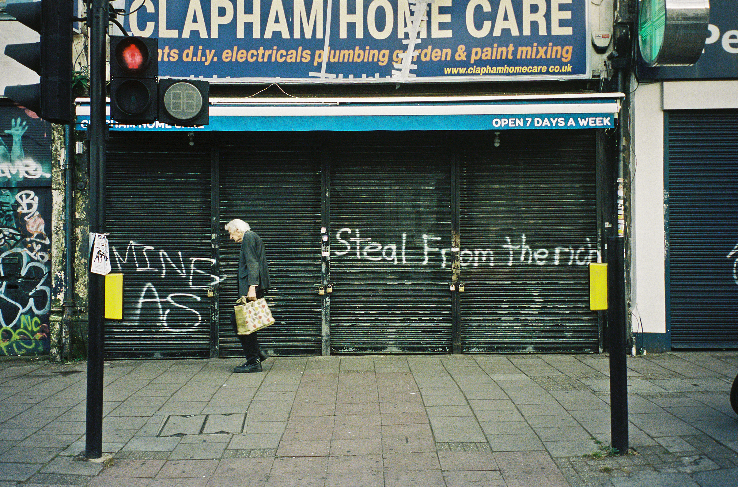Colour photograph of an elderly person walking along the street past a shuttered shop front that has “STEAL FROM THE RICH” spray-painted in front