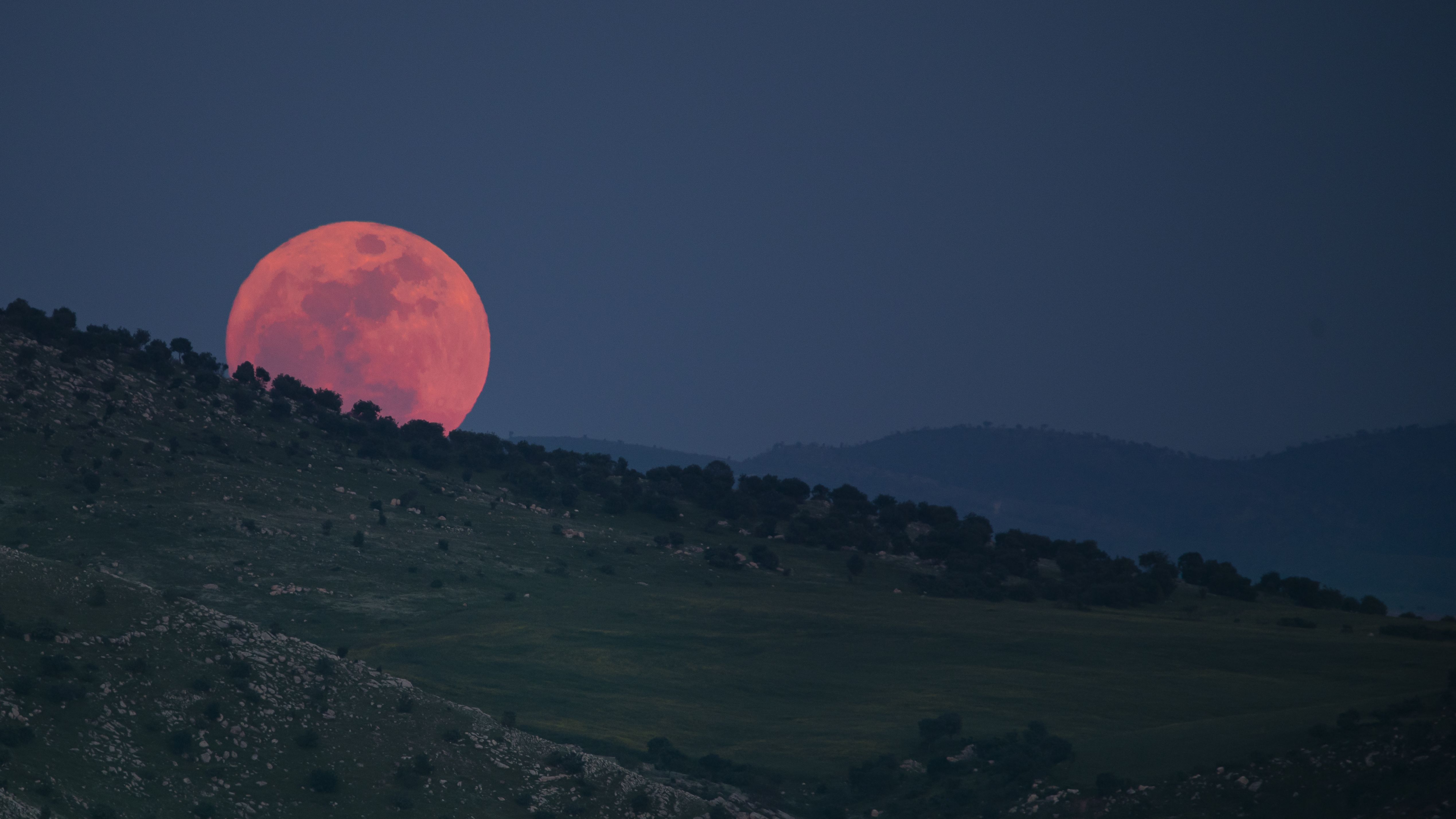 A red full moon rises behind a hillside at night.
