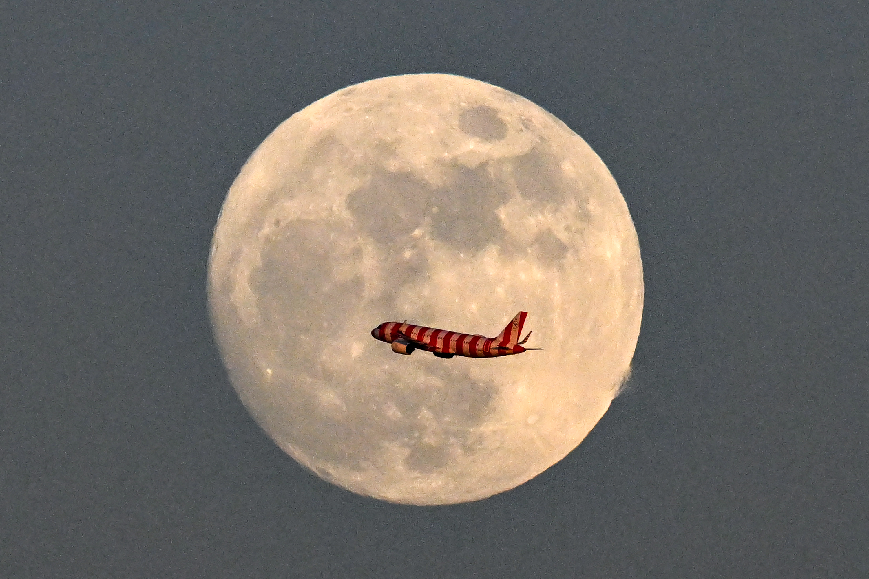 A passenger jet is photographed flying across the face of a full moon in the grey evening sky.
