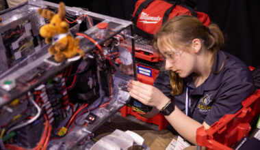 A student holds onto the edge of the rectangular body of a prototype lunar robot as she works on it. The rover has a rectangular body, which has multicolored attached to it. There is a small stuffed moose sitting on top of the rover.