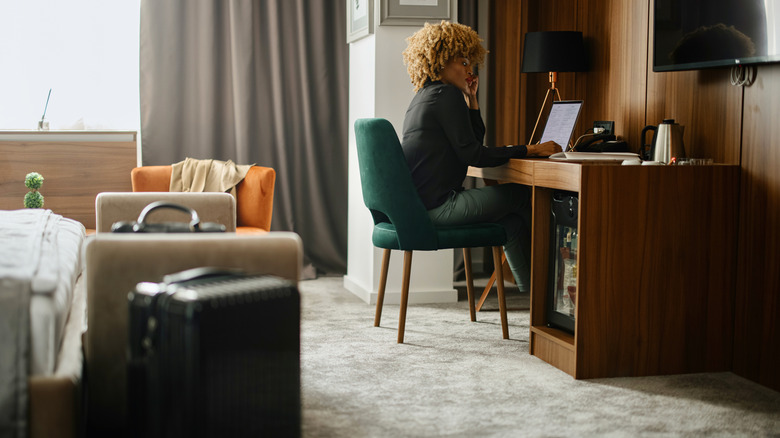 A woman sitting at a desk in a hotel on her laptop, suitcases by the bed in the foreground.