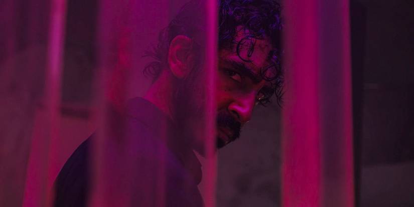 Kid (Dev Patel) looking furious, seen through sheer curtains, illuminated by neon light in Monkey Man
