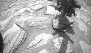 A black and white still of NASA's Curiosity Mars rover just lifting its drill arm with a rock stuck to it