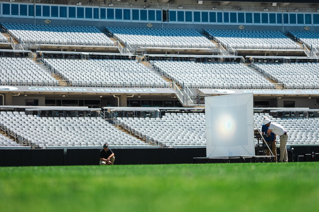 a large white square stands on a football field, supported by braces securring it to a platform. People stand around. A bright central light hits the center of the square.