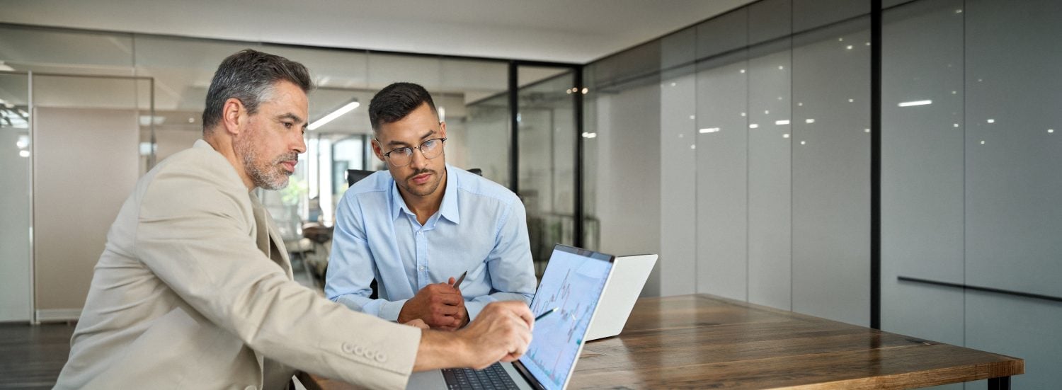 Two busy business men investors doing financial trading market data analysis. Male advisor and analyst consulting about stock exchange investing pointing at chart on laptop computer working in office.