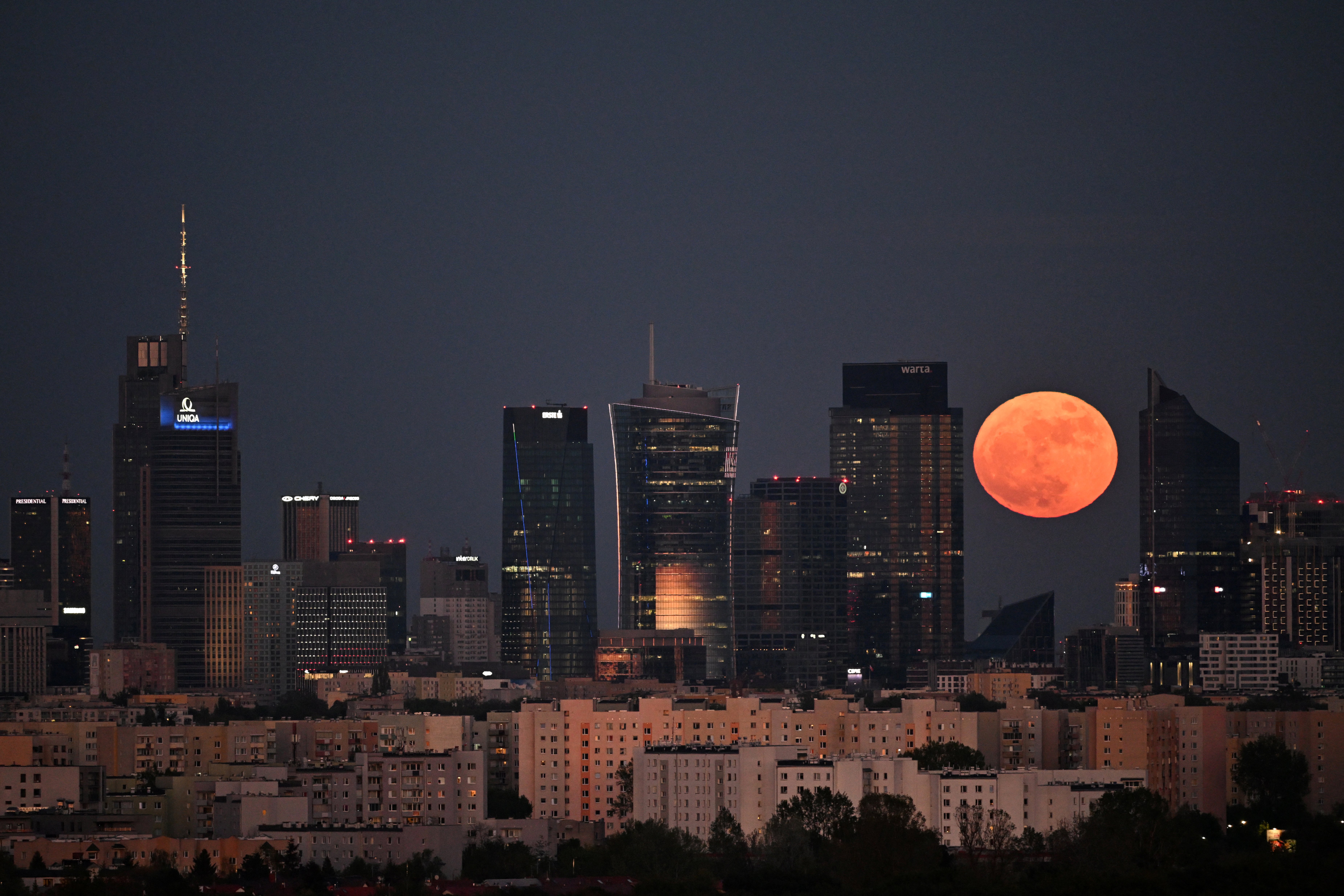 An orange full moon is pictured rising to the right of a city skyline photo at night between skyscrapers.