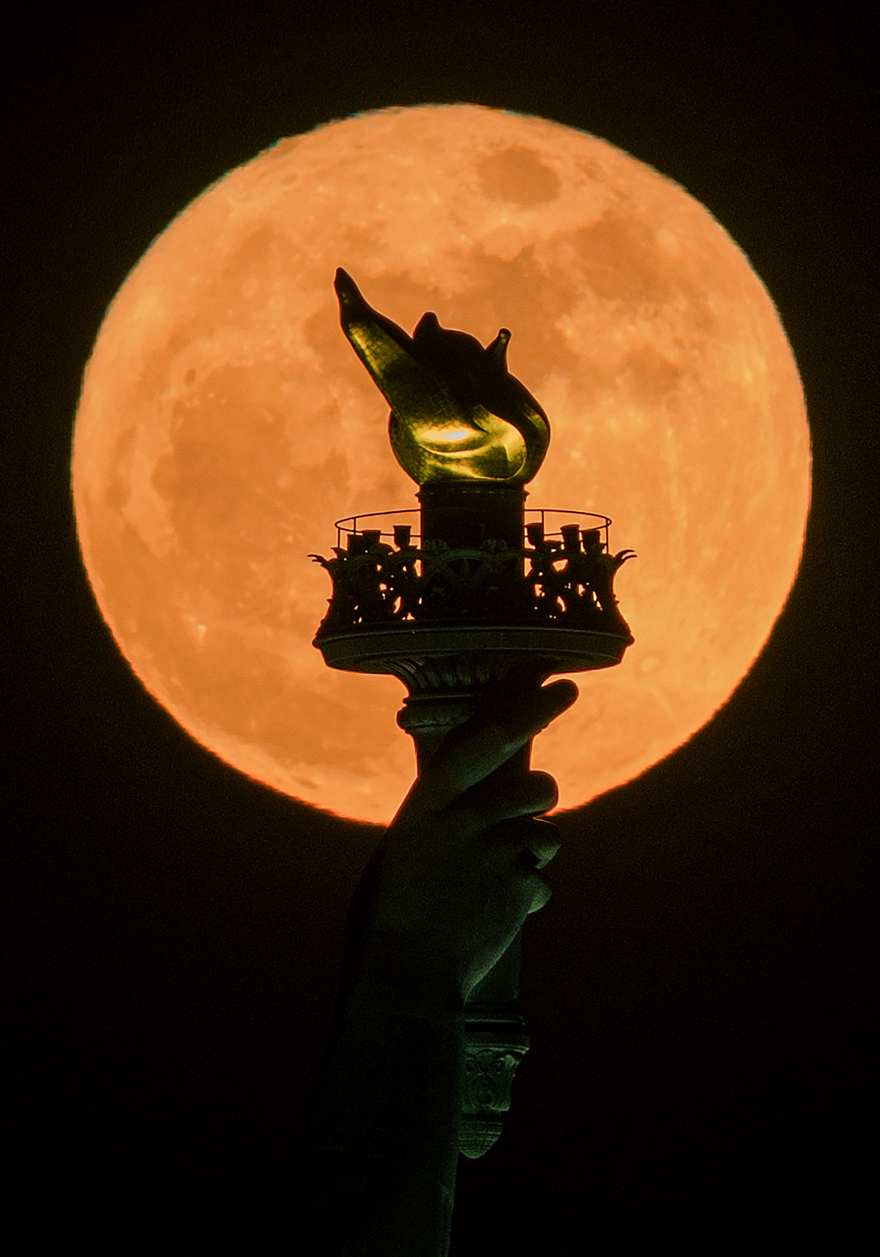 A yellow-orange full moon is pictured rising behind the silhouetted torch of the Statue of Liberty at night.