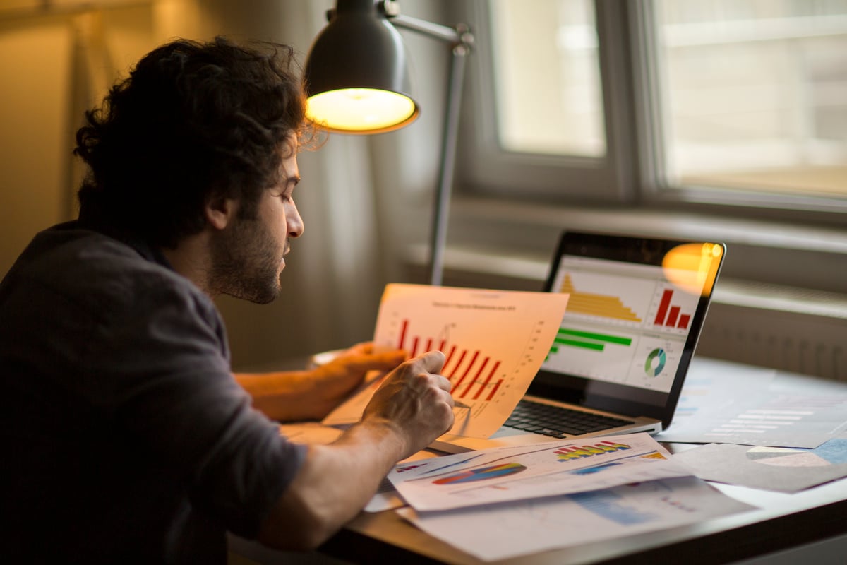 An investor holding a chart, comparing the data to numbers on a laptop.