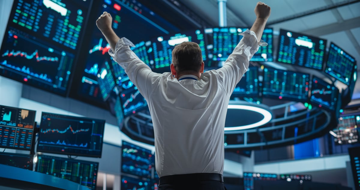 A person facing away from the viewer raises their fists in triumph as they watch a set of stock price monitors.