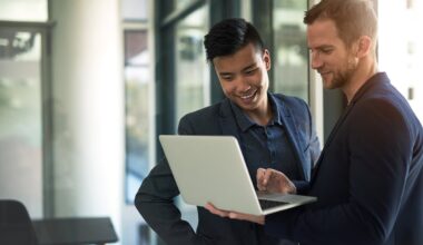 Two investors, standing in an office, look at something on a laptop.