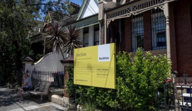 A Ray White auction sign stands outside a row of terrace houses in Sydney, with a person seated on a bench nearby.
