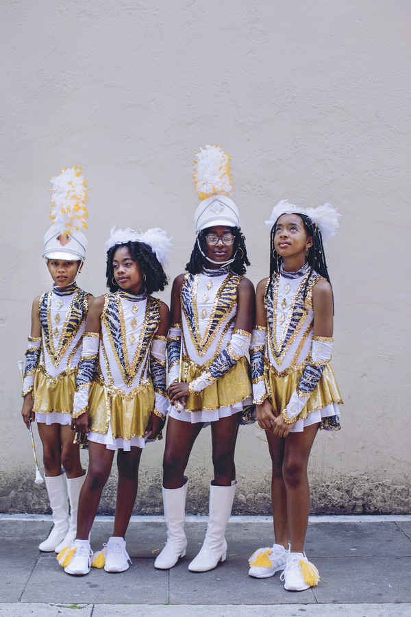 Four young Black girls in matching gold and white sequinned majorette uniforms and tall feathered headdresses stand against a plain wall, each looking in a different direction