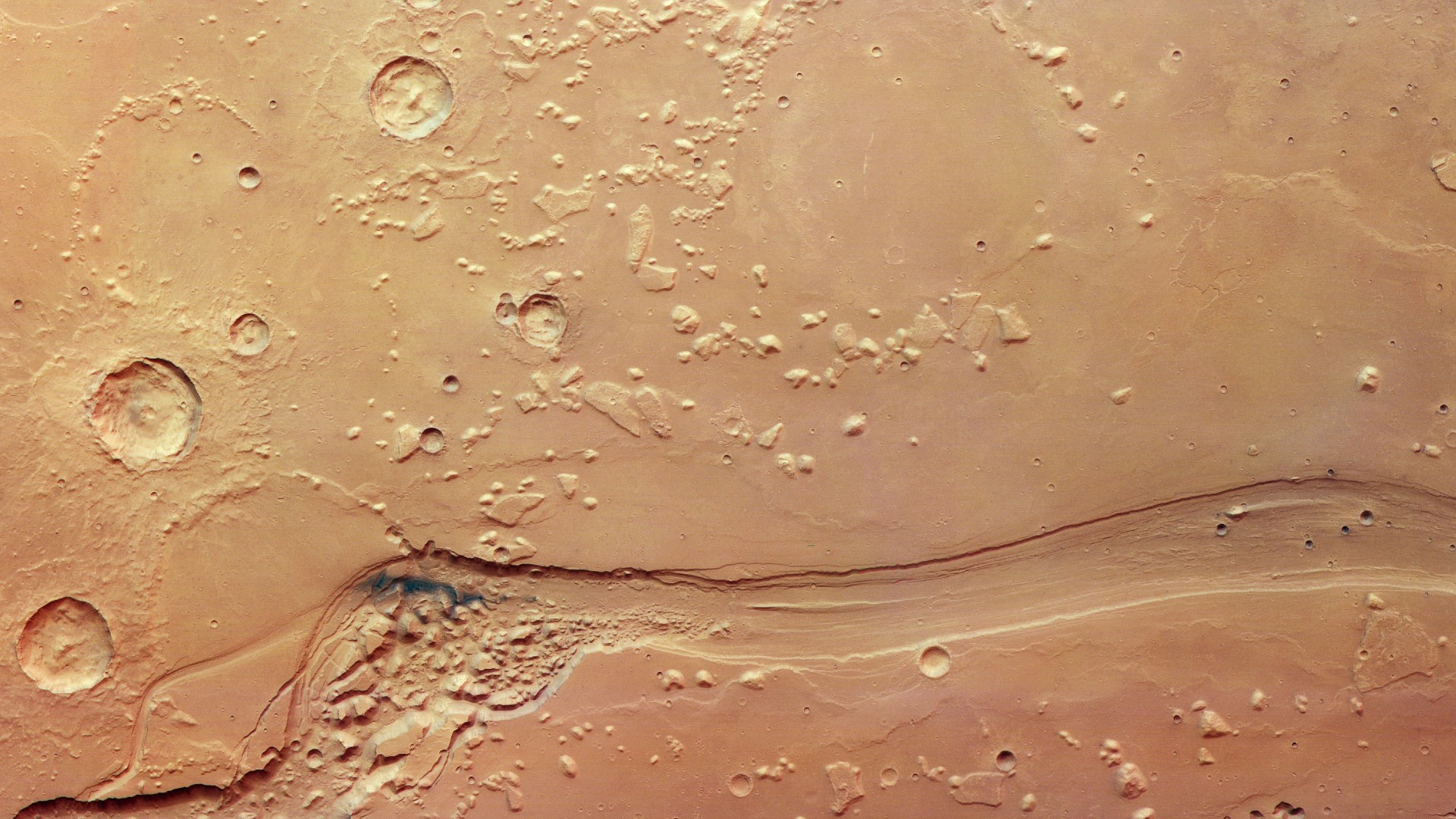Looking down on a patch of Mars's surface, with a winding canyon carving from the left to the right of the frame. On the left, the canyon widens and is filled with lumpy, knobbly terrain. On the right, the canyon is more straight and streamlined. Surrounding the canyon is more lumpy terrain, but on top of a smoother surface