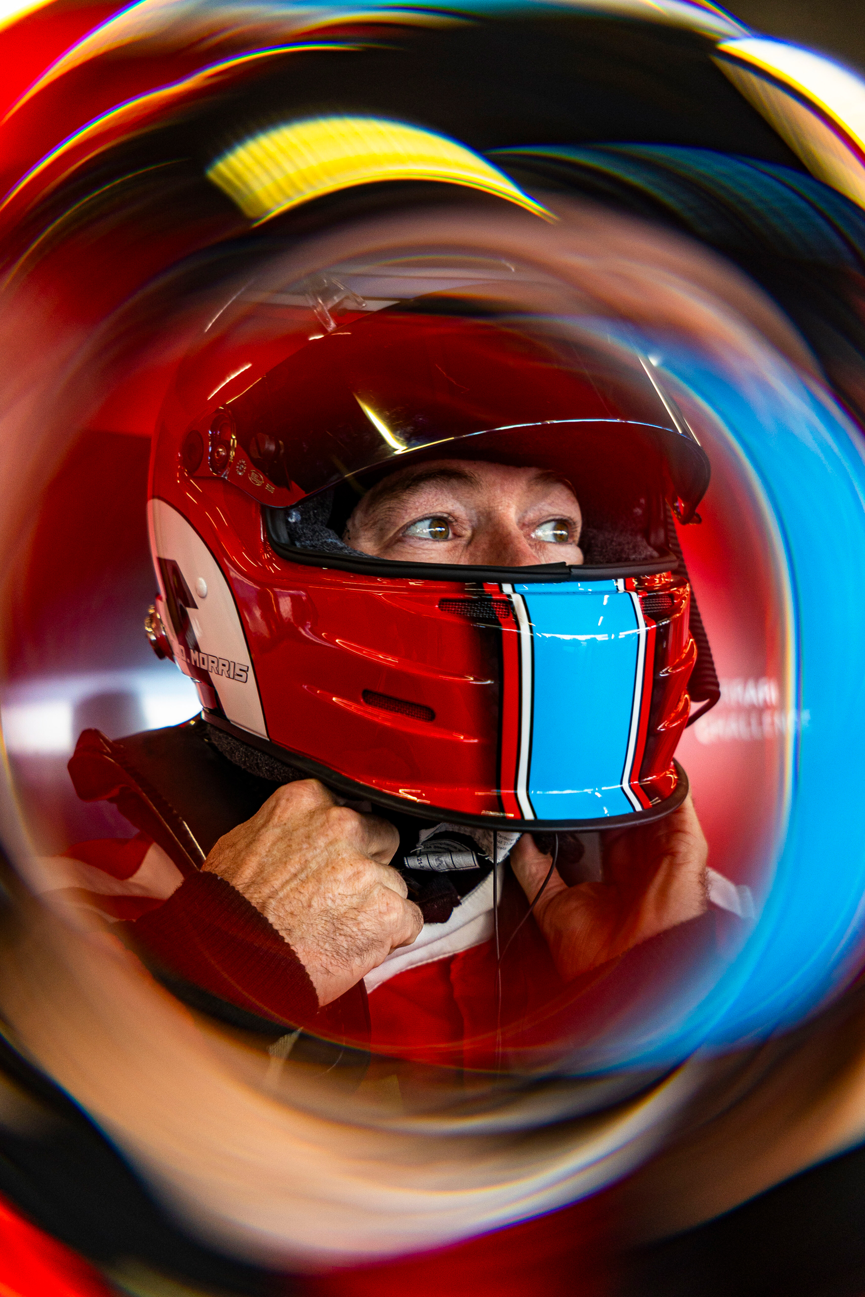 Race car driver in a red helmet fastening the chin strap, with a swirling motion blur effect around the image