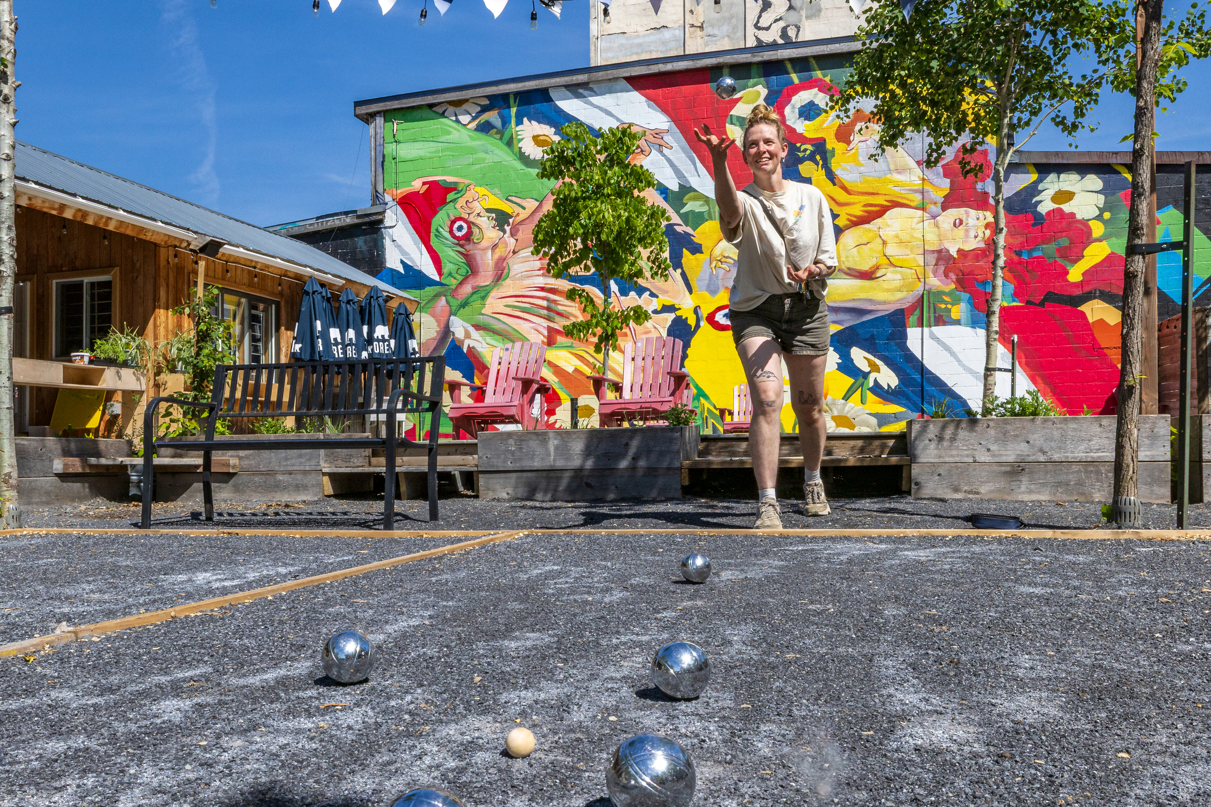 A woman launches a pétanque bar in the air on an outdoor court on a patio.
