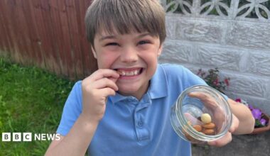 A young boy wearing a blue polo-shirt, smiling with a bowl of nuts in his left hand. He's also holding a nut up to his face.
