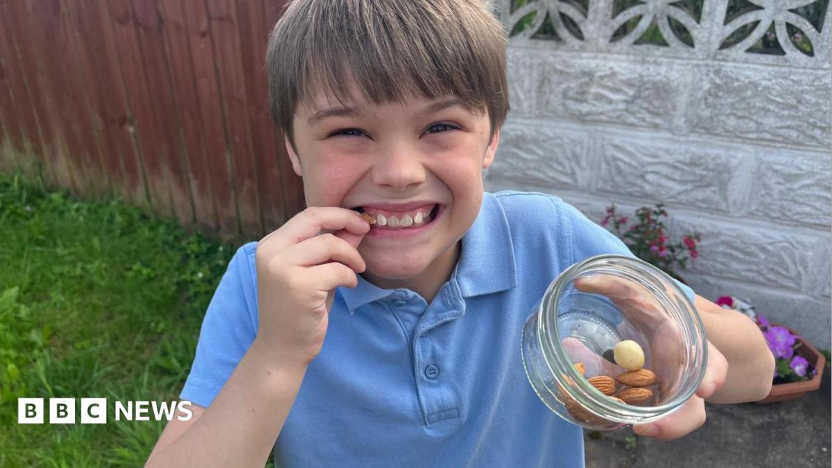 A young boy wearing a blue polo-shirt, smiling with a bowl of nuts in his left hand. He's also holding a nut up to his face.