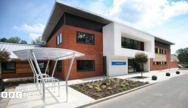 The front of the St Aubyn Centre. It is a red brick building with a cycle rack outside. There are rainbow-coloured windows to the side and it has a few plants in a flowerbed at the front.