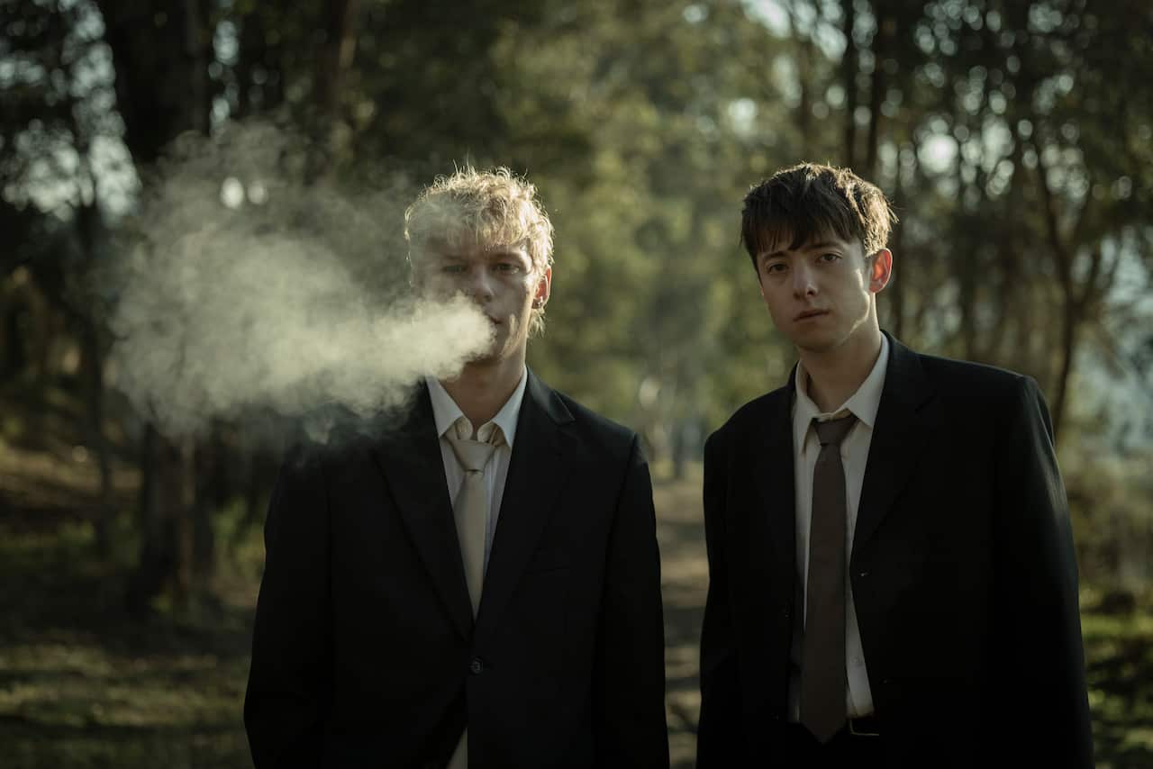 Two teenage boys wearing suits standing in the outdoors. 