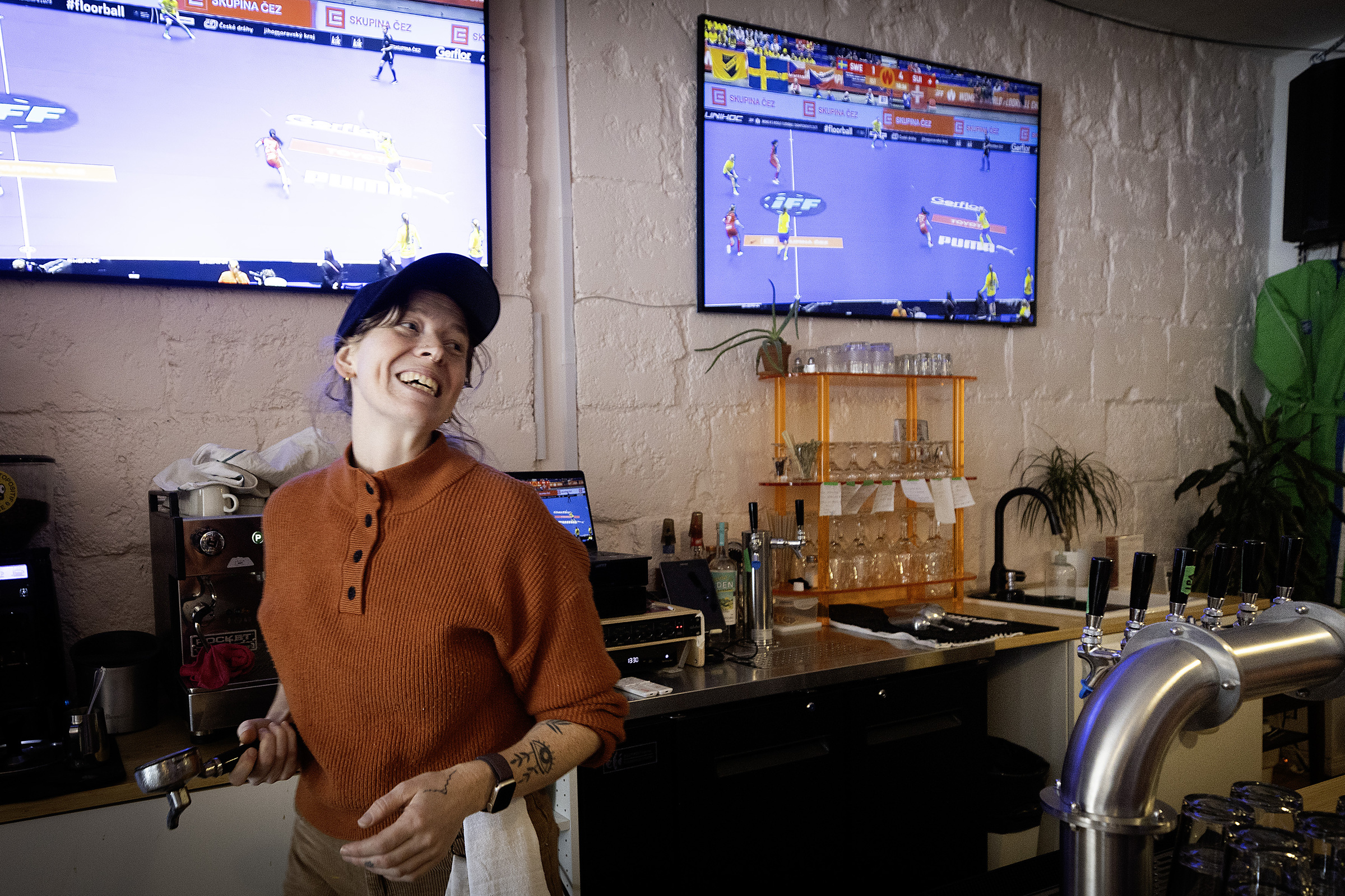 A woman smiles as she handles a coffee machine behind the bar, with TV screens showing a game behind her.