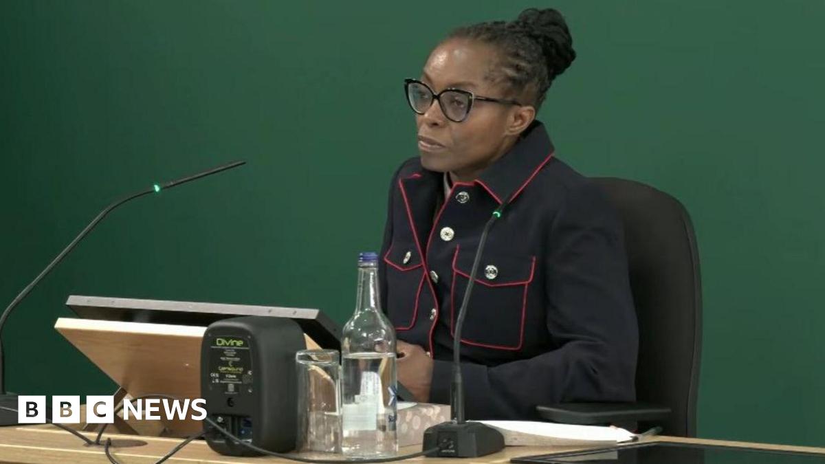 A woman sat a desk in a blue jacket with red trim in front of a green back drop.