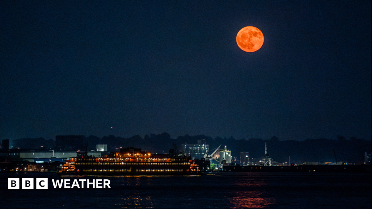 An orange-hued moon hangs in the sky over a river illuminated by lights from nearby properties