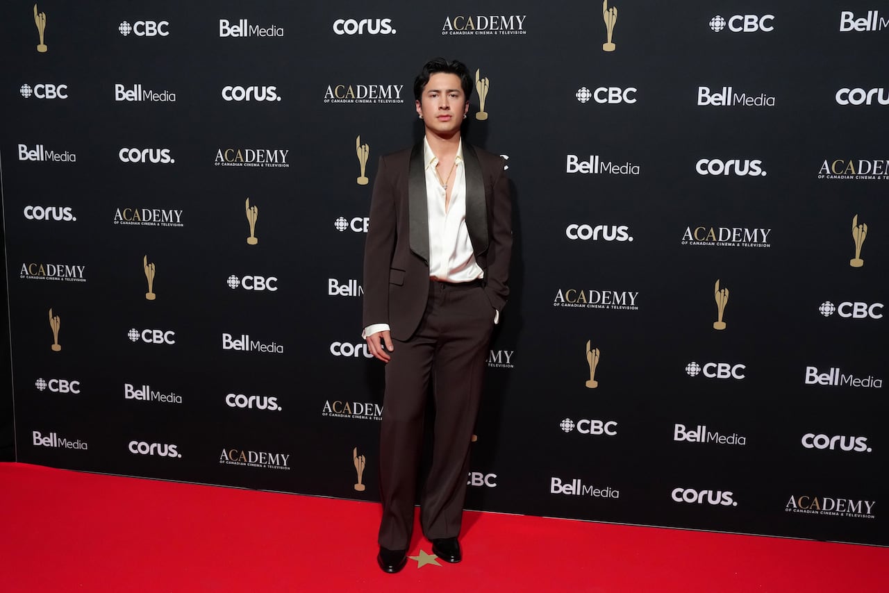 A man poses for a photo in a brown tux and a white shirt with a few buttons undone and no tie.