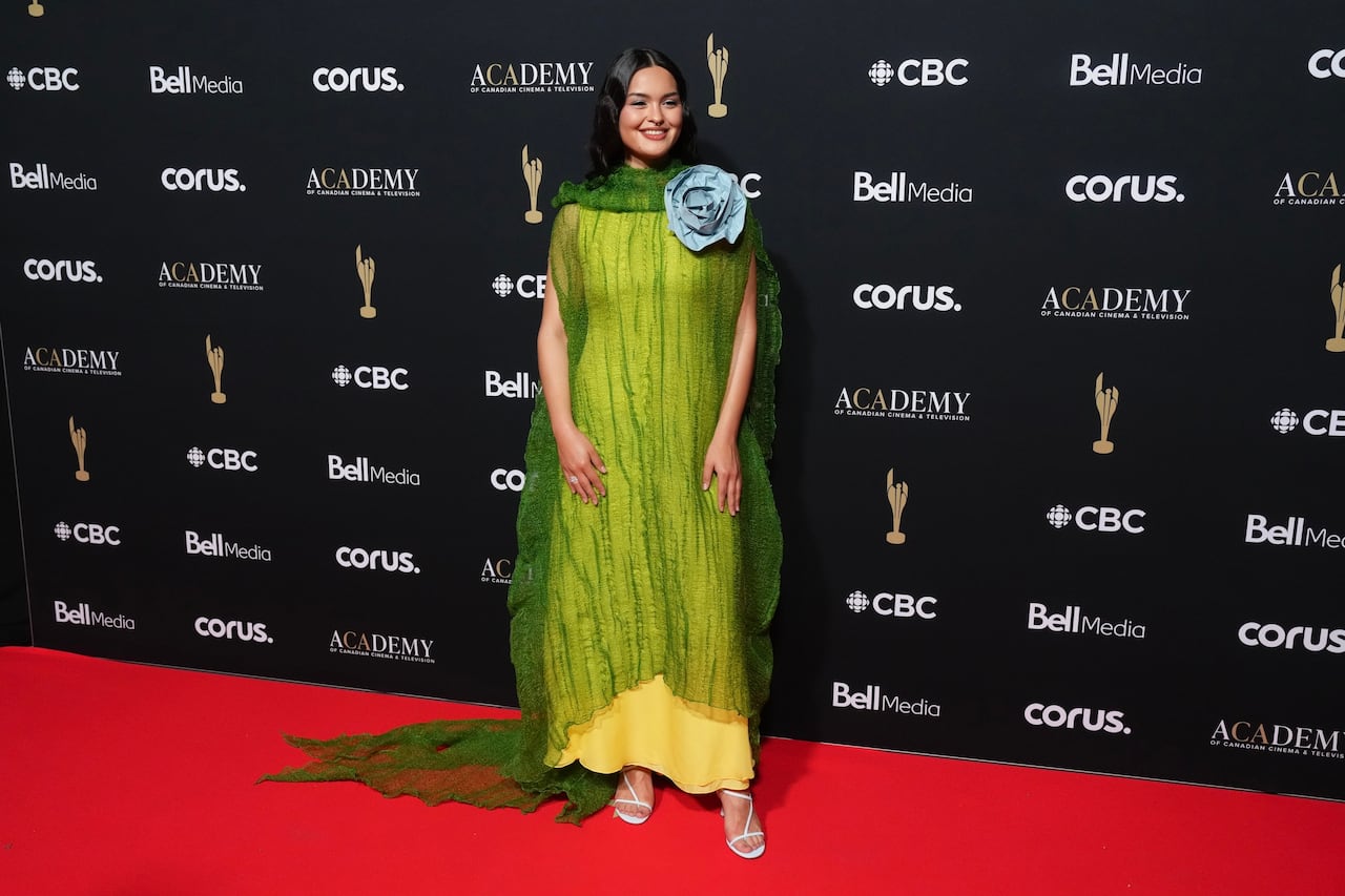 A woman poses for a photo wearing a yellow dress with a sheer green layer over it and a blue flower at the shoulder.