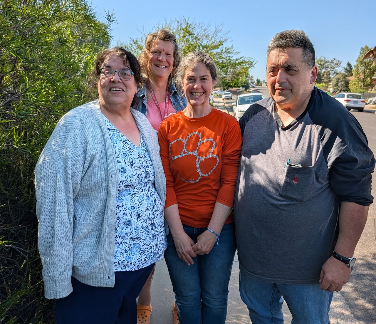 A woman with dark hair and glasses, wearing a grey sweater, black pants and a floral shirt, stands to the left of two other women and one man on the right, on a narrow paved sidewalk in front of trees.