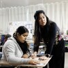 Mary Acebu, a Filipino woman wearing all black with a flowered vest and knitted beret, leans over a desk to help a student at her desk. The student has long brown hair in a ponytail and a gray sweatshirt.