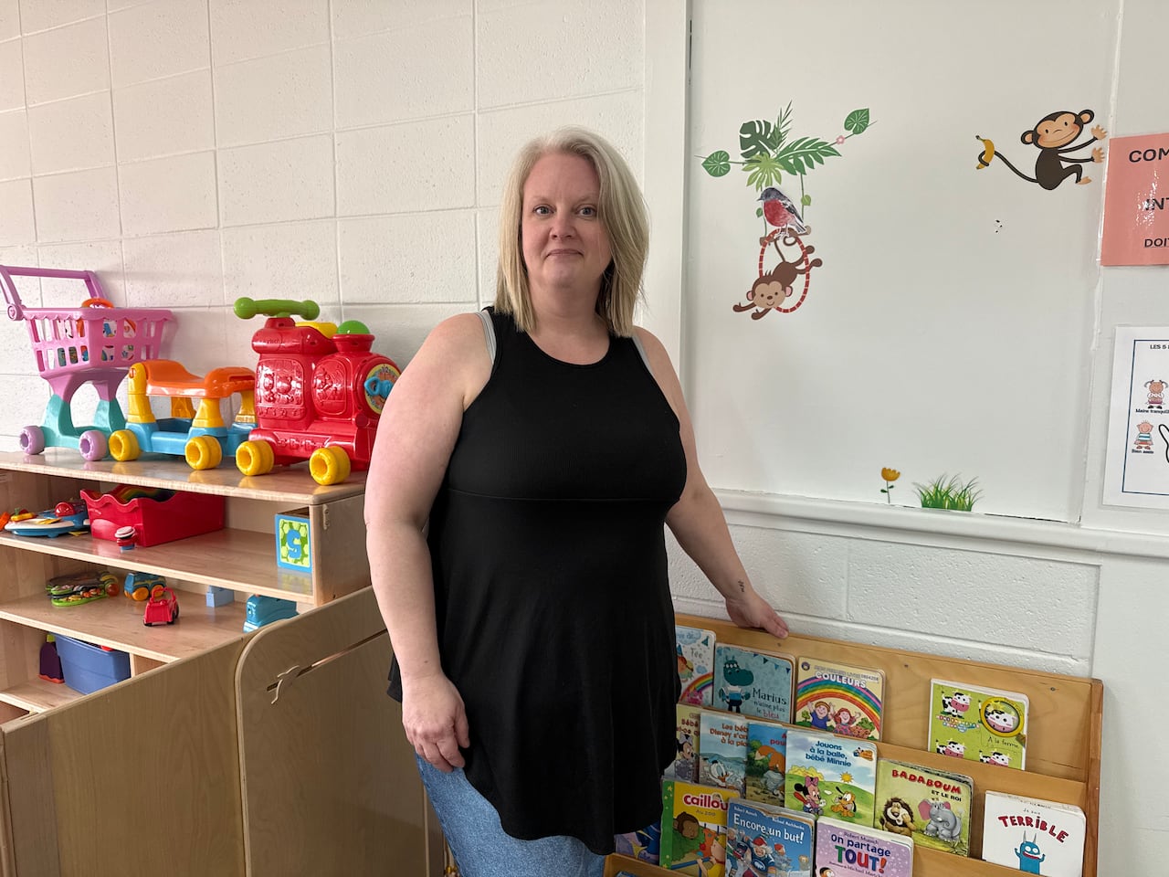 A woman wearing a black tank top and jeans stands in front a children's book shelf in a daycare centre.