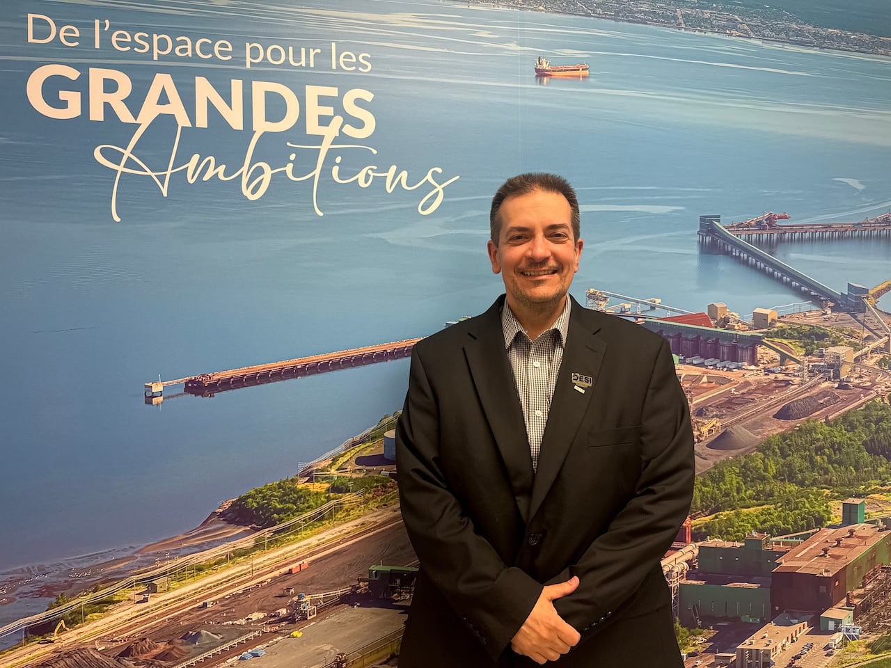 A man in a suit stands in front of a mural depicting the Port of Sept-Îles, with a caption in the top left corner saying: "De l'espace pour les grandes ambitions"