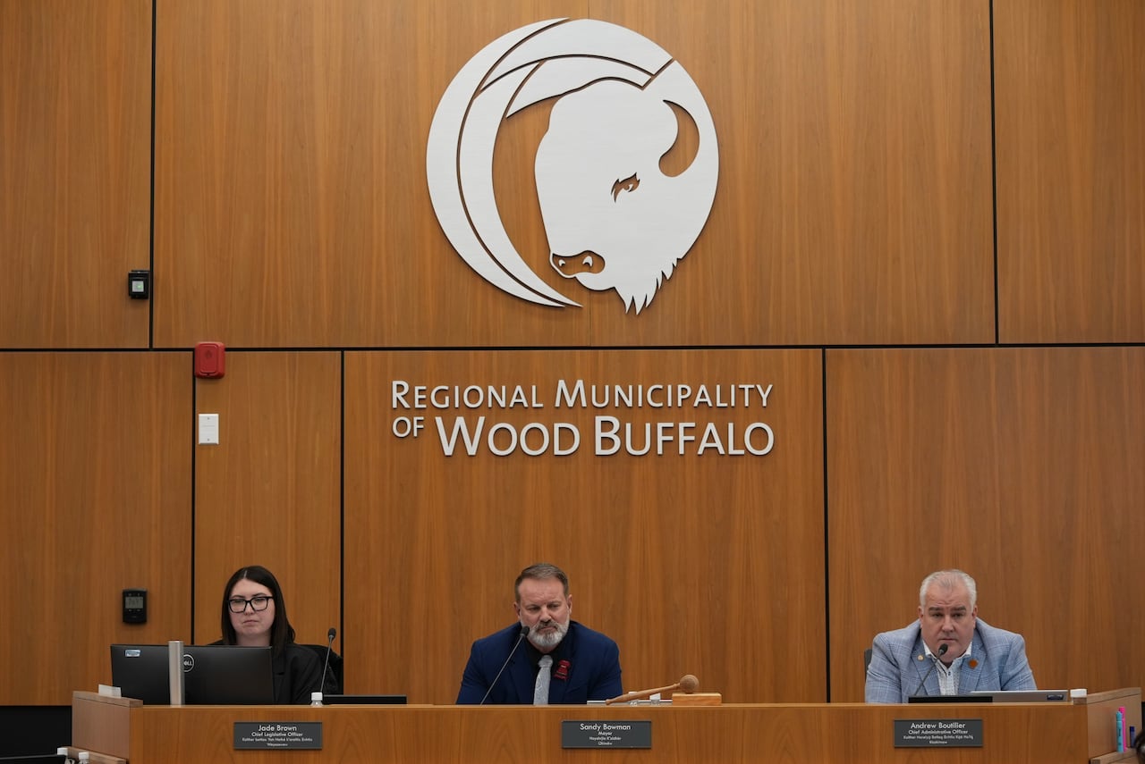 Three municipal employees sit at a wooden desk in front of a wood-panelled wall and beneath a large image of a bison's head and the words "Regional Municipality of Wood Buffalo."