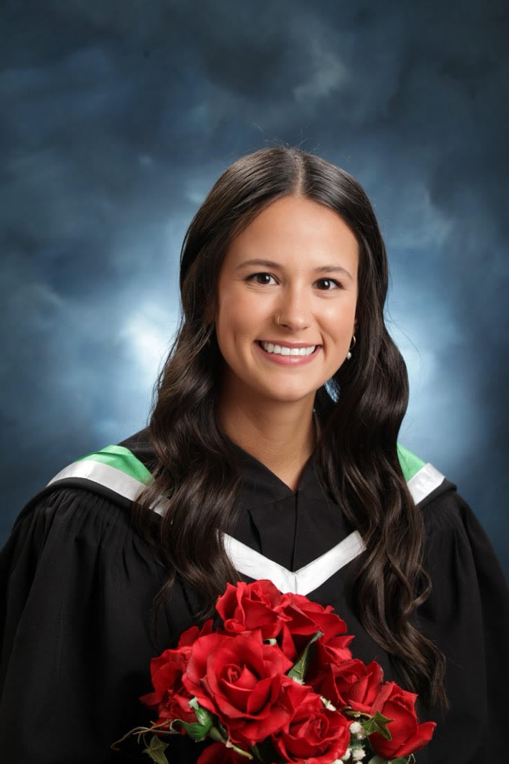 A young woman with long black hair smiles and wears a graduate's gown while holding a bouquet of red roses.