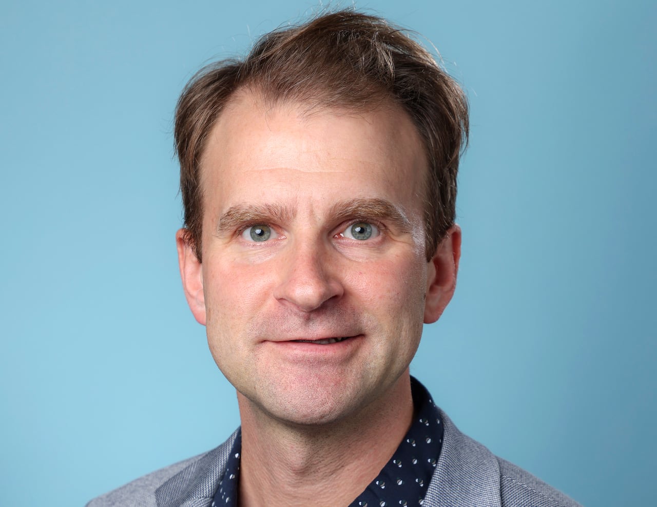 A man with brown hair, wearing a navy dress shirt and grey blazer, smiles for a headshot.