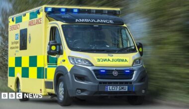 An East of England Ambulance Service ambulance driving on a road. Next to it is a row of green bushes which are blurred, giving the impression that the vehicle is moving fast.