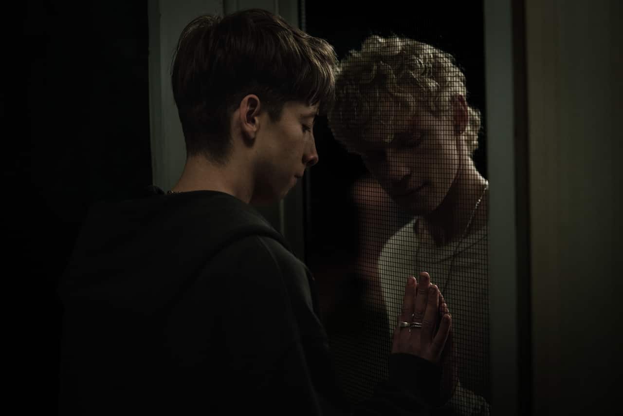 Two men touch each other's hands through a screen door.