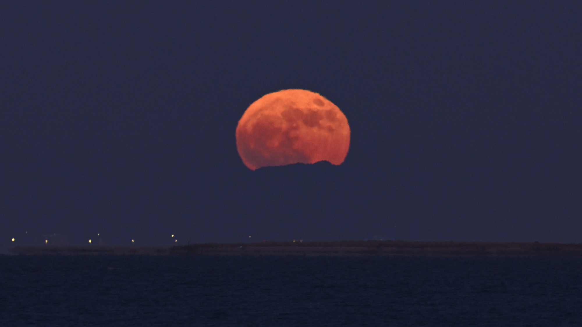 a large strawberry red moon rises over a large body of water.