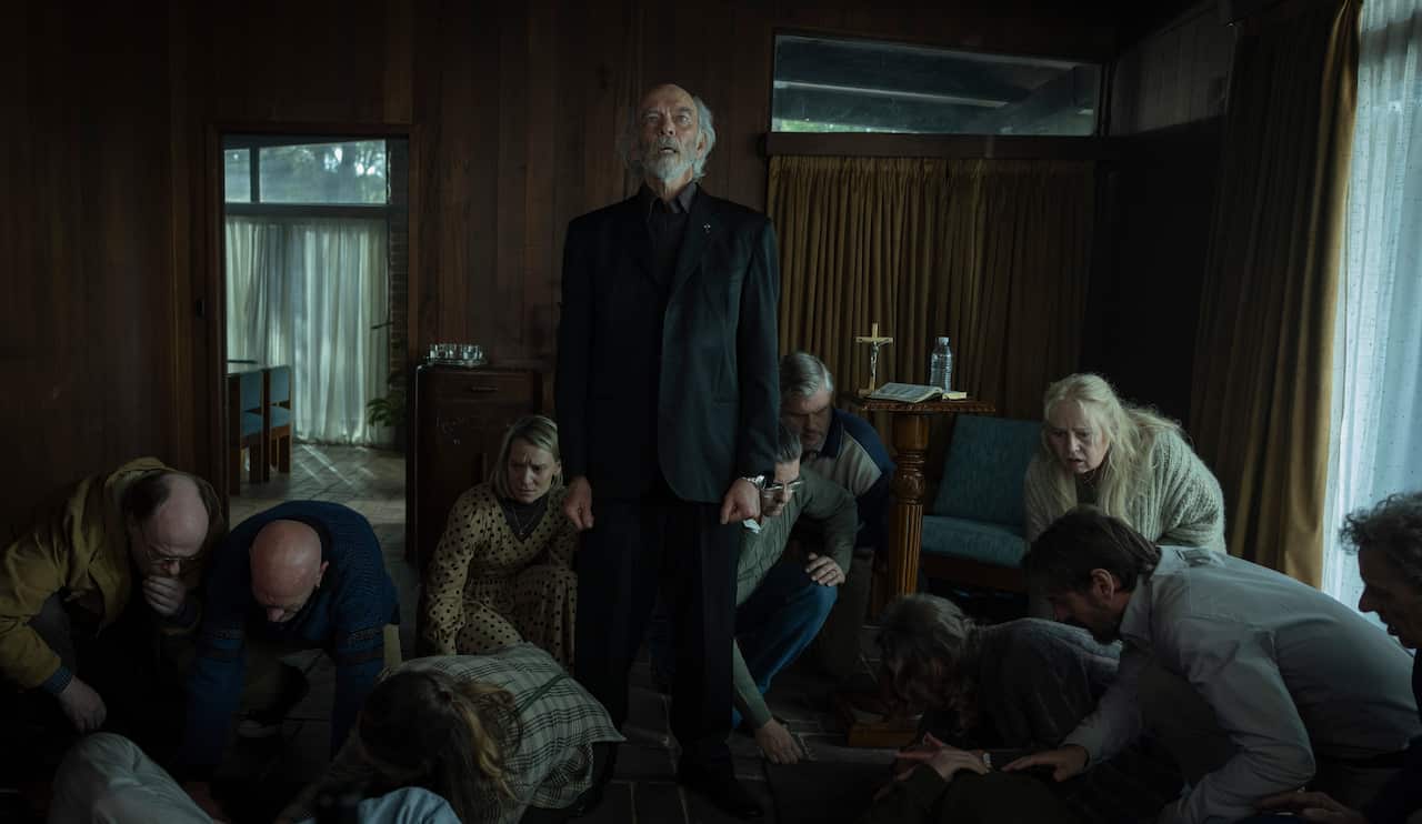 A priest dressed in a black suit stands in a dark room while people huddle on the floor around him.