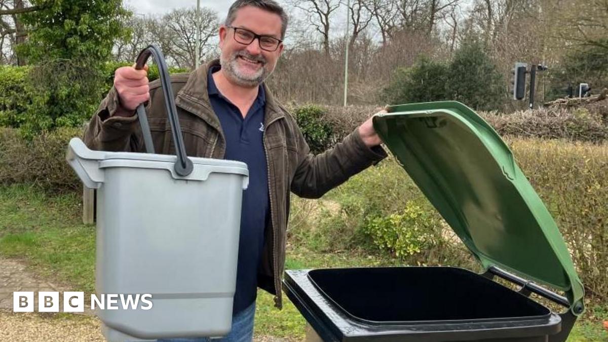 Jon Neal is standing on a gravel drive with trees and bushes in the background. He is holding a small grey caddy in one hand while standing next to a black and green wheelie bin, which he is holding open. He is wearing blue jeans, a blue jumper and a brown jacket. He is wearing glasses.