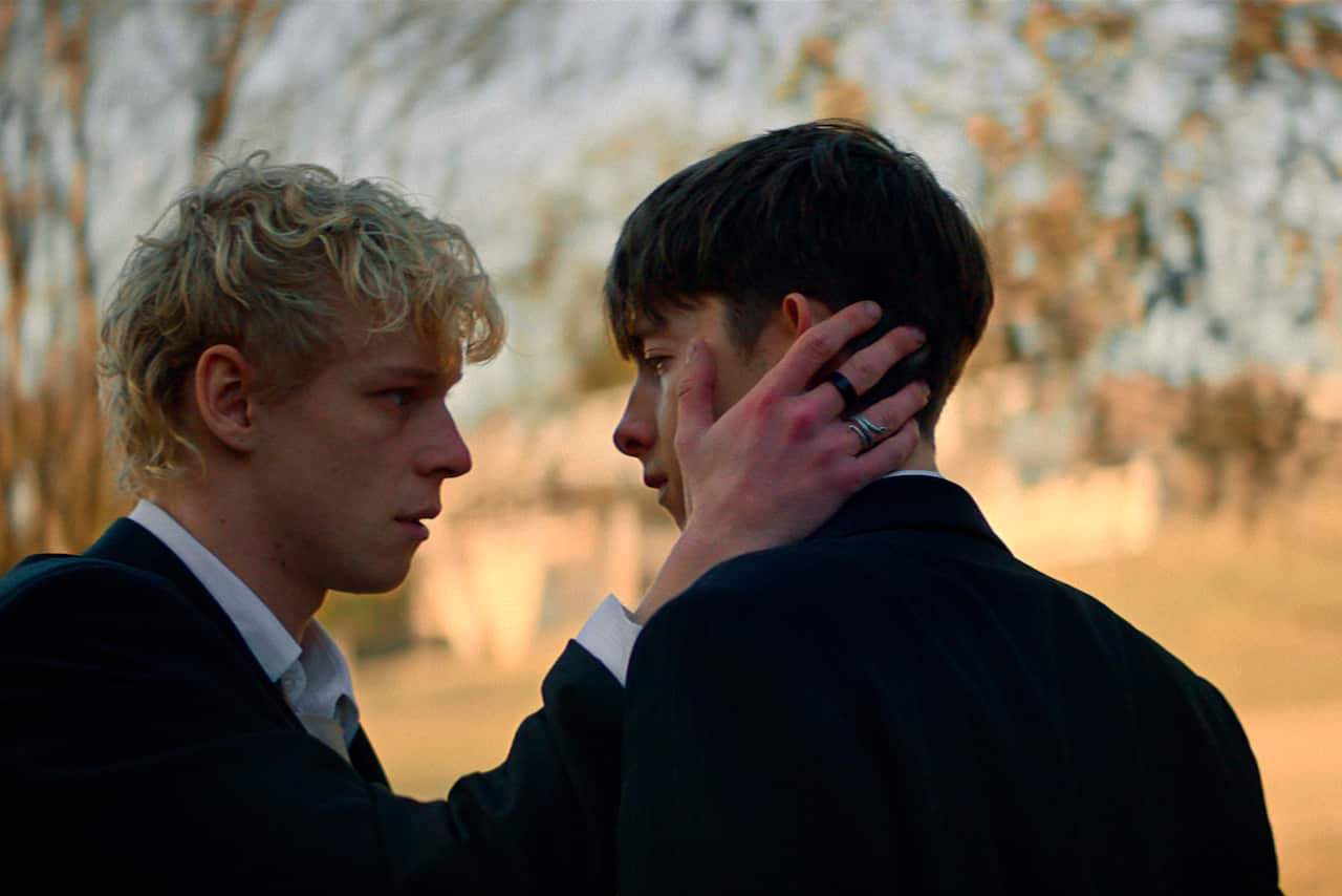 A blonde teenager grabbing the face of a darker-haired teenager. They are outside and are both wearing suits.