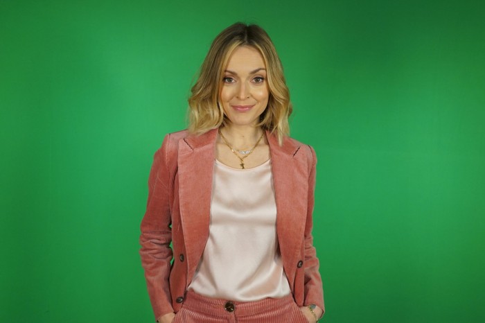 409440,TITLE:Radio Presenters Fearne Cotton wearing a pink pantsuit with a silk blouse, smiling ahead in front of a green background.