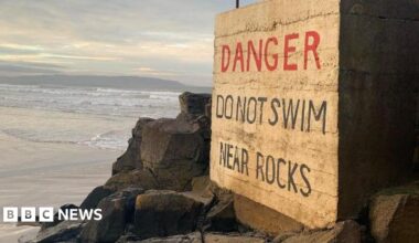 Portstewart Strand. A large sign reads: 'Danger – Do Not Swim Near Rocks.' It is positioned beside a wide area of jagged black rocks, with the shoreline visible in the background.